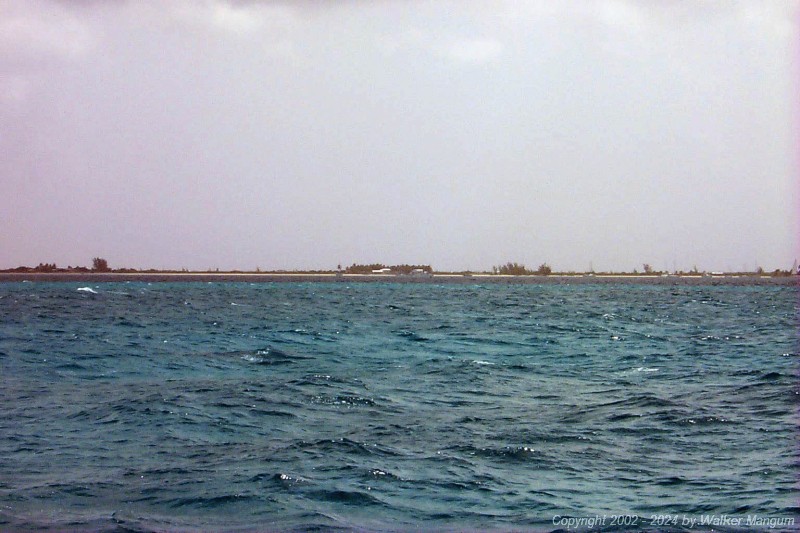 Approaching Anegada. It is slightly hazy and we are 1 mile from the entrance buoys. You can see Setting Point Villa, the Australian pines at Setting Point Villa, the palm trees at Neptune's Treasure, the white roof of Neptune's Treasure, the boat house at Neptune's Treasure, the red entrance buoy, the Australian pines at Whistling Pines, and the Lobster Trap Restaurant.