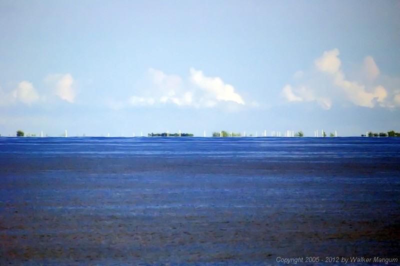 Anegada from 12 miles away, photographed from the top of the ferry with a long telephoto lens. Sailboats in the anchorage are "hull down", with only the tops of their masts visible above the horizon. The buildings ashore are below the horizon.